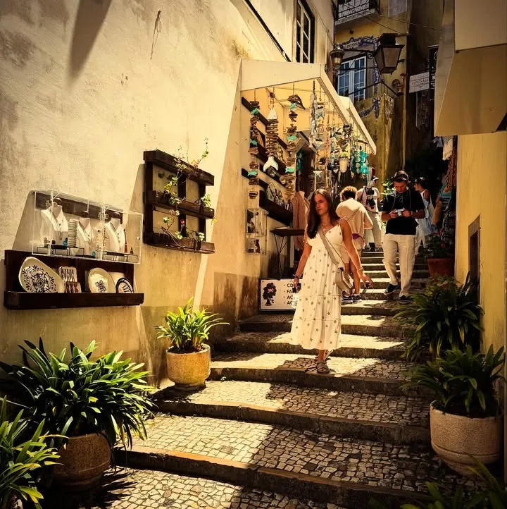 A woman in a white dress descending a sunlit stepped alley lined with shops and plants