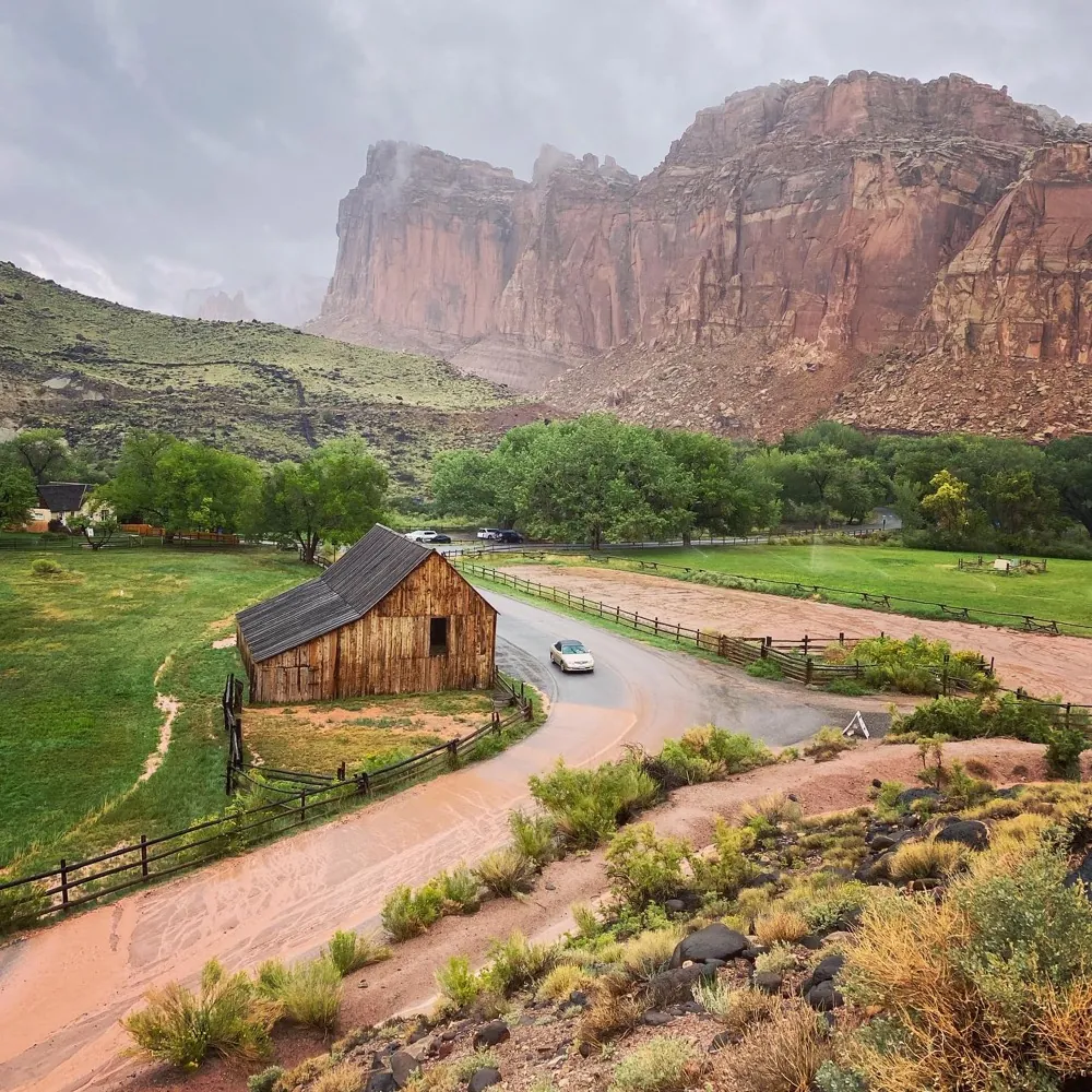 A wooden barn on a green valley floor beneath towering red cliffs