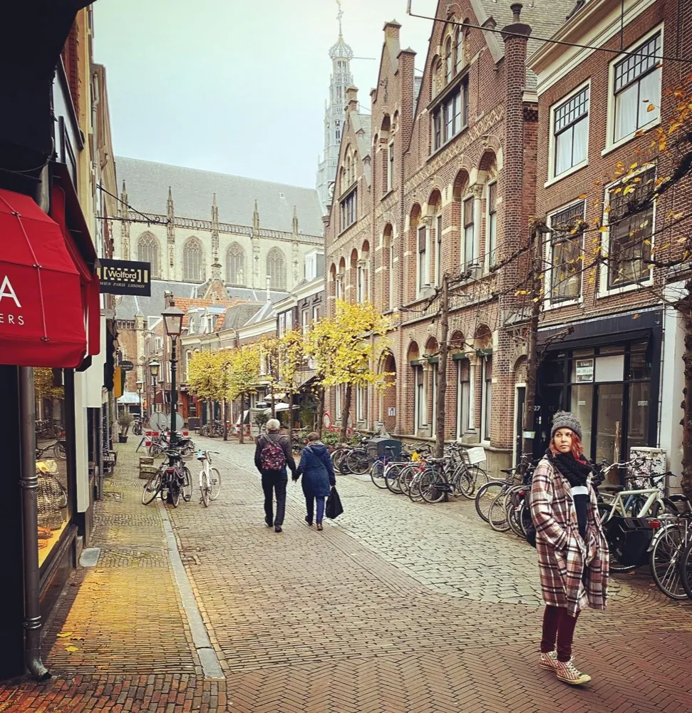 A woman in a plaid coat glancing back on a cobbled street lined with bicycles and autumn trees