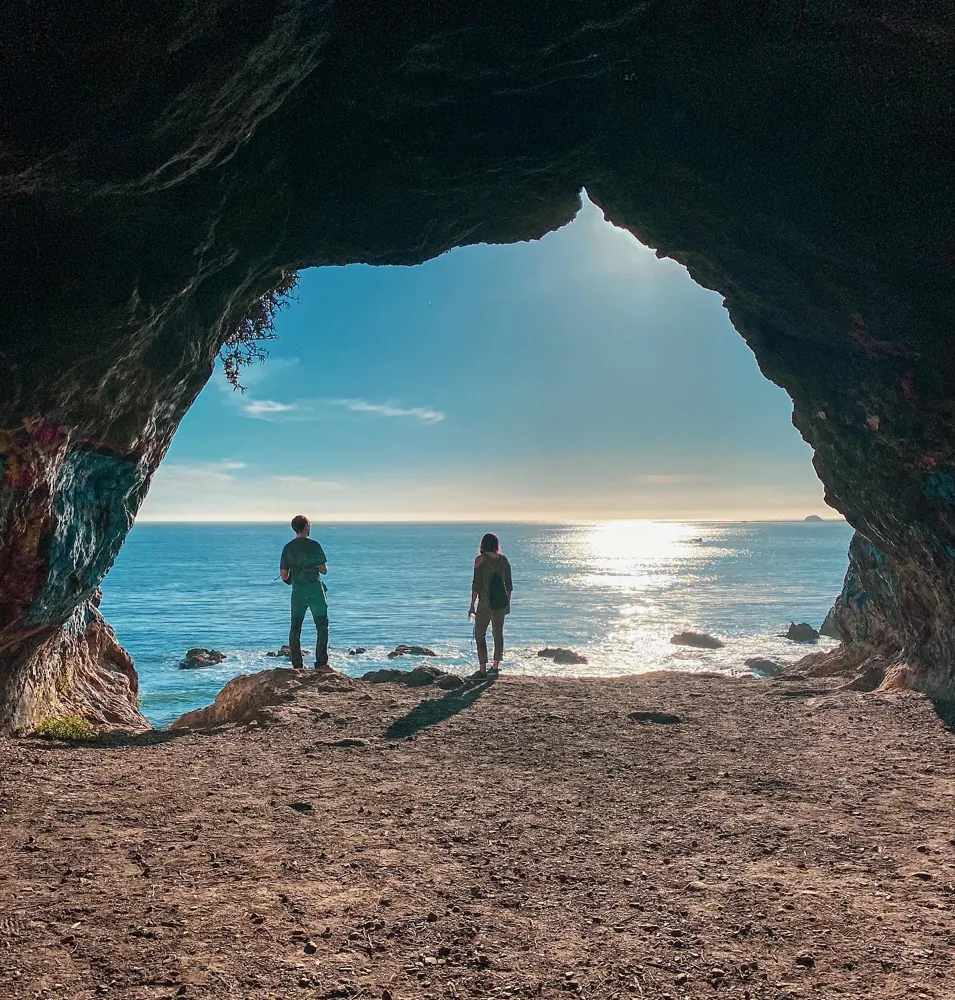Two people silhouetted at the mouth of a sea cave above a sunlit ocean