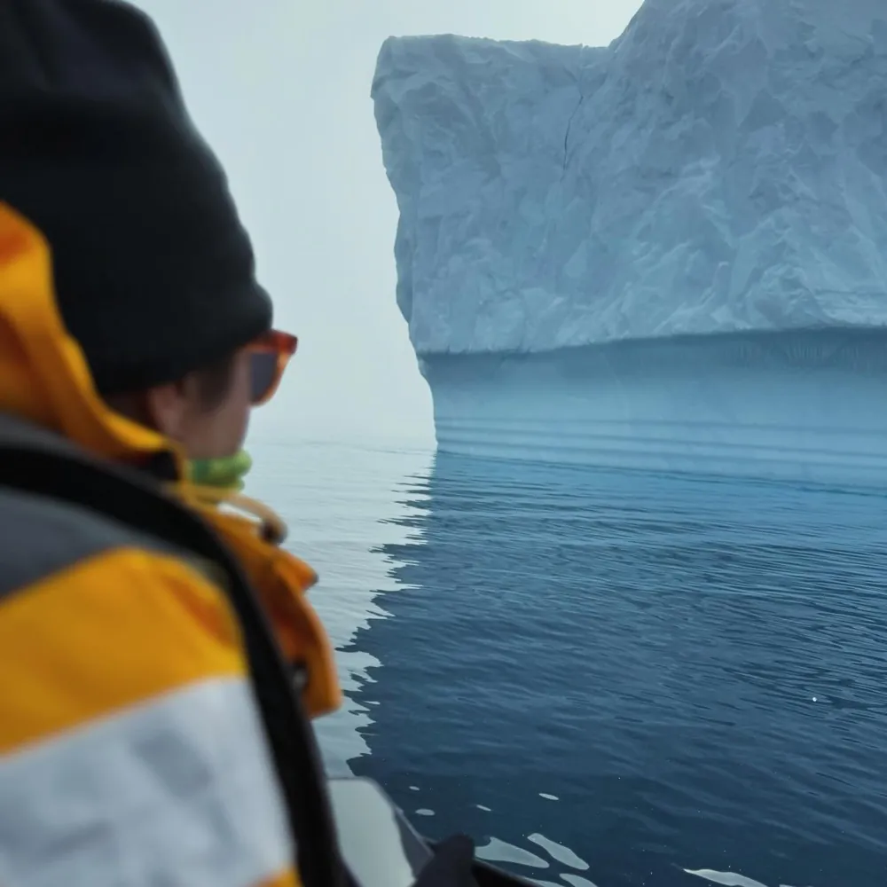 A person in an orange parka watching a flat-topped iceberg from a small boat on still water