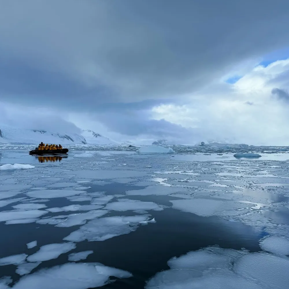A boat of people in yellow parkas crossing broken pack ice under a heavy sky
